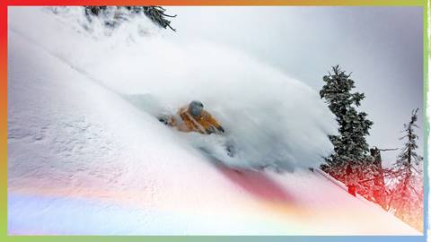 A snowboarder in a yellow jacket and black helmet slashing through a section of untouched powder after a winter storm.