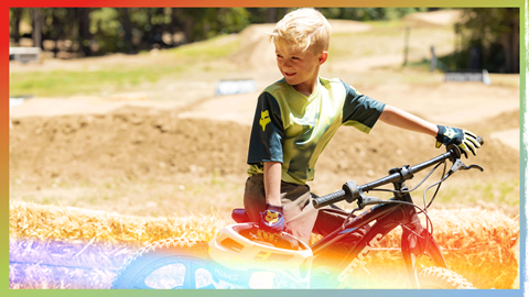 Kiddo with blonde hair, in a green color jersey, sitting on mountain bike and holding a full face helmet with Smith branded goggles attached to it.