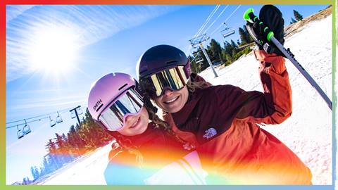 Adult, in red jacket with black helmet and snow goggles holding hand up with ski pole, and a kiddo, in dark jacket with pink helmet and snow goggles, on the winter slopes during a sunny day.
