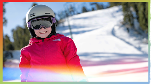 child snowboarder wearing a pink jacket with a helmet at goggles smiling at the camera with snow summit behind her