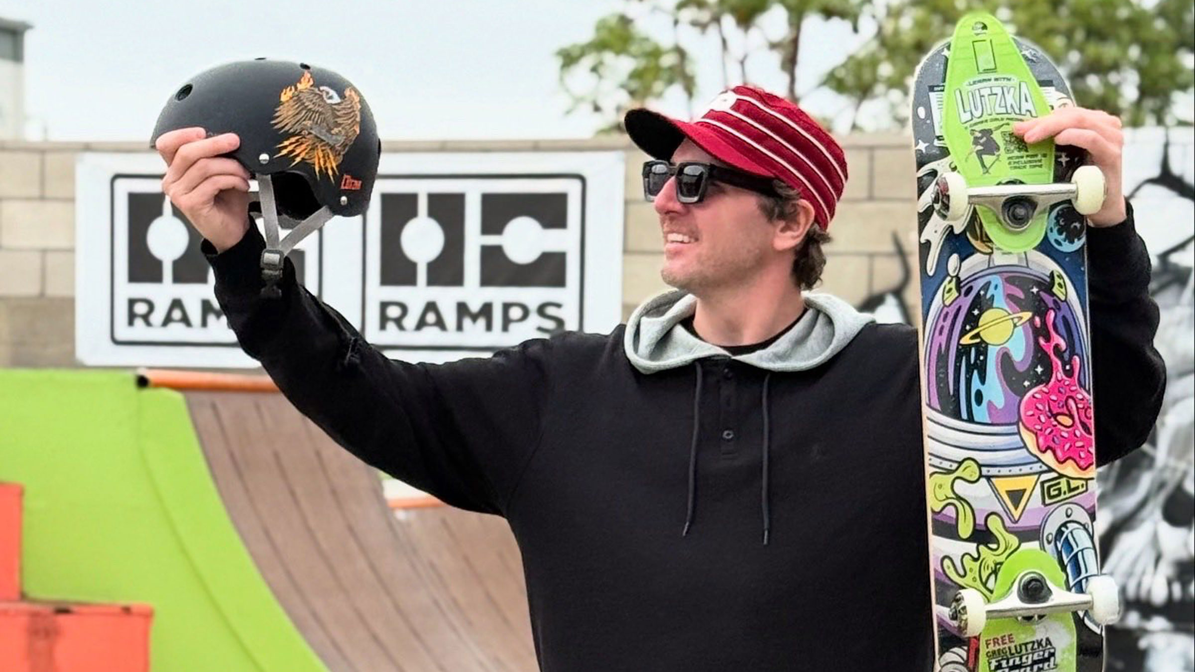 Pro skater, Greg Lutzka, in a red had and black hooded sweatshirt in front of an OC Ramp mini halfpipe holding a helmet in one hand and skateboard in the other hand.