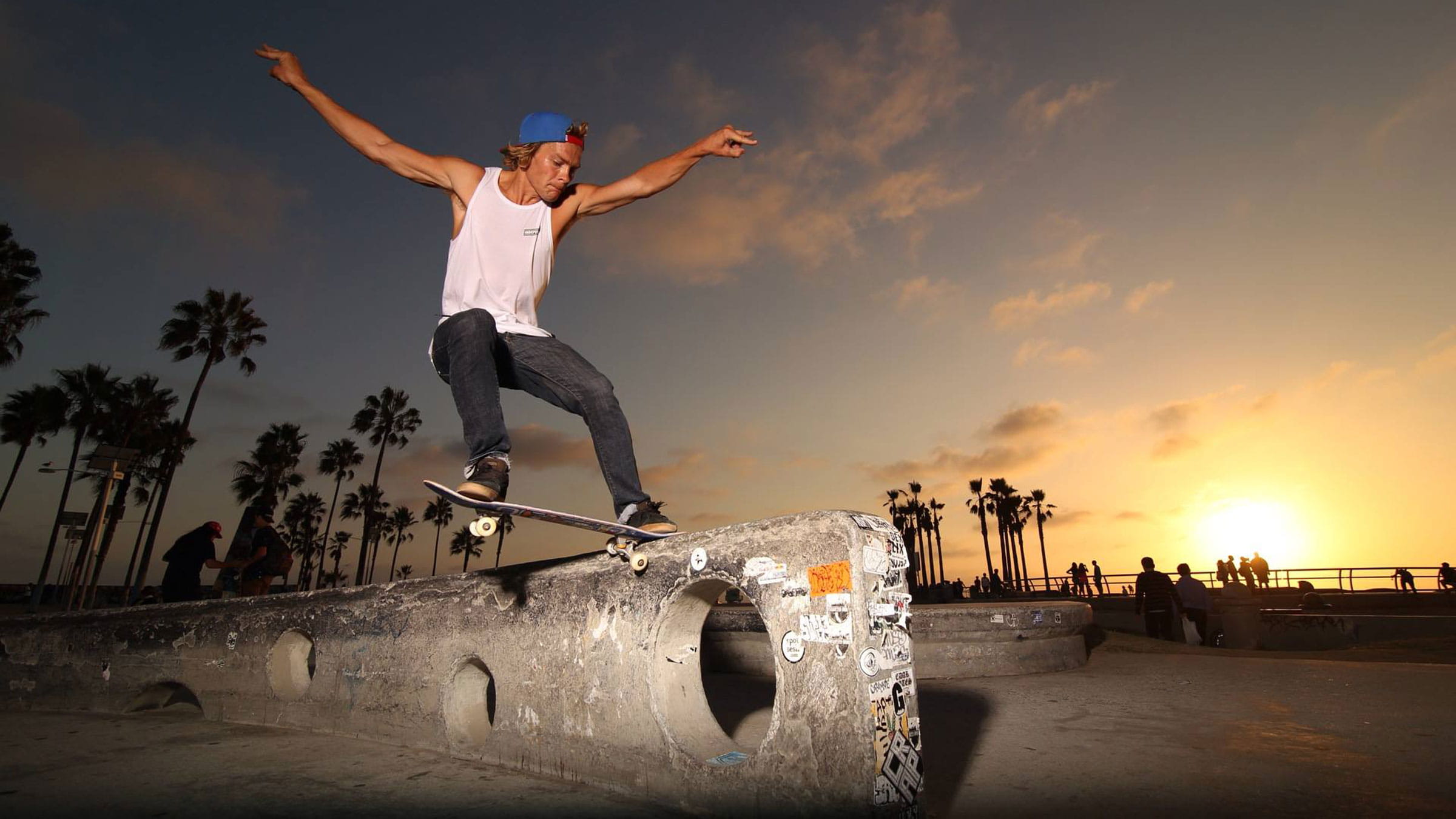 Pro skater, TomTom Ryen, wearing dark pants and a white tank top doing a tailslide grind on skateboard on a ledge by the beach during sunset.