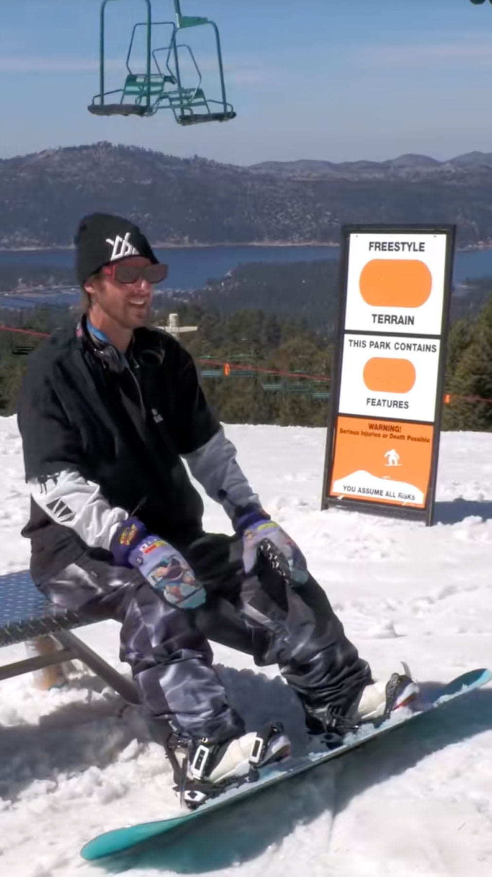 Snowboarder in black top, beanie, and gray snow pants, strapped onto their snowboard sitting on a bench at the top of a Snow Summit trail leading into the terrain park and overlooking Big Bear Lake, California.