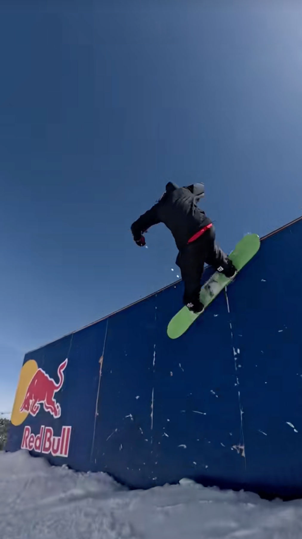 Snowboarder with a green board and wearing all black, riding on the outside of the Red Bull Ride-Thru container jib located on the slopes of Bear Mountain.