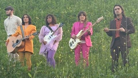 A five person band, standing in a high grassy field, consisting of three females dressed in bright pastel suits playing their guitars and two men without instruments looking off into the distance.