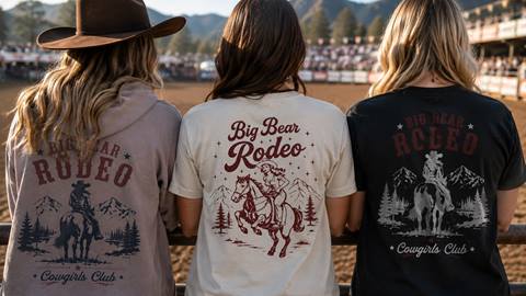 Three women with long hair with their backs turned to the camera, looking at a arena rodeo ring, while wearing Big Bear Rodeo merchandise.