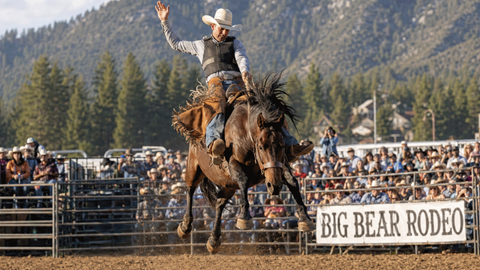 A cowboy riding a horse during a rodeo event in an outdoor arena with a Big Bear Rodeo sign.
