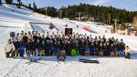 24-25 Team Bear group photo with the athletes and coaches standing on the snow at the base of Bear Mountain on a sunny winter day.