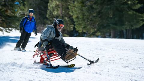 An adaptive skier using a mono-ski carving down a snow trail with a USARC guide in blue jacket following closely behind.