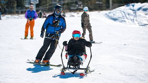 An adaptive skier in black snow outerwear and red helmet using a bi-ski and carving down a snow trail with a USARC guide in blue jacket following closely behind.
