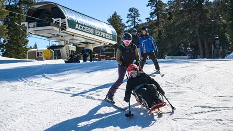 Adaptive skier with a guide being pushed in a ski chair and moving down a Bear Mountain trail near the Access Express chairlift on a sunny blue sky day.