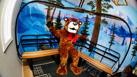 Biggie the Bear, Big Bear Mountain Resort's mascot, sitting on a makeshift chairlift bench with a painted mountain landscape mural on the wall inside Biggie's Den at Snow Summit
