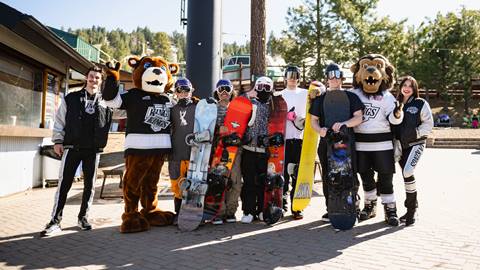 Big Bear Mountain Resort mascot, Biggie, standing in the Snow Summit base area with the Los Angeles Kings hockey mascot and two employees, plus five snowboarders holding their gear, on a sunny winter day.