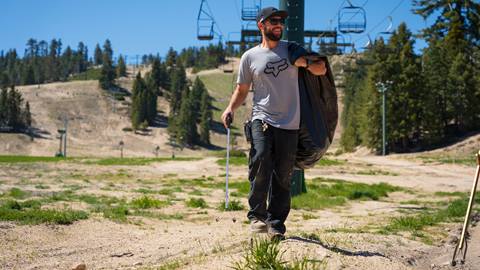 Sunny blue sky day at Snow Valley with ad adult in dark pants and a gray shirt on the summer trails, under a chairlift, holding a trash bag over one shoulder.
