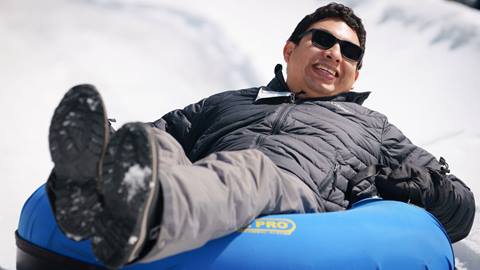 Adult in a black snow vest with grey pants riding in a snow tube while zooming down a lane at Snow Valley's Coyote Creek Tube Park
