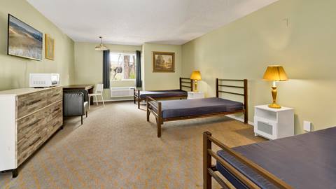 An inside view of an employee housing room with three beds, a dresser, nightstands, desk, lounge chair, and window that looks into the forest.
