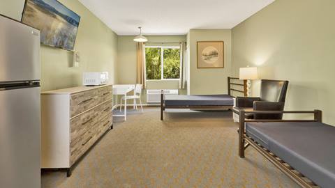 Employee housing room with two beds, a nightstand, comfortable lounge chair, dresser, microwave, mini fridge, and desk that is placed next to a window.