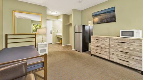 Employee housing room looking towards the entrance door with a bed, a nightstand, dresser, microwave, mini fridge, and open closet to hang clothes.