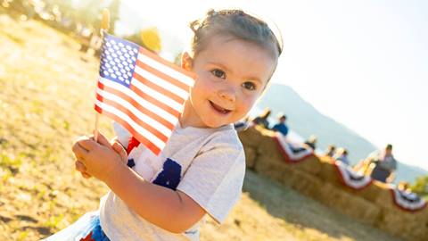 A smiling kiddo in a white shirt holding an American flag at the top of Snow Summit for the annual Fourth of July Above the Boom firework event.