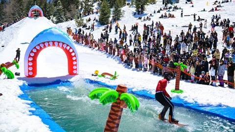 A snowboarder in a red jacket with afro wig helmet riding through the pond skim at Bear Break 2024