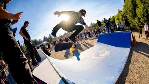 A skateboarding doing a mid-air trick on a blue wood mini halfpipe with a crowd watching.