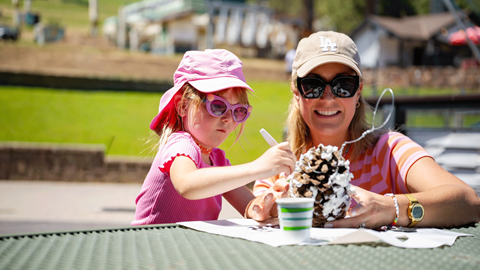 Young kid wearing a pink hat and shirt making a pinecone bird feeder with her mom