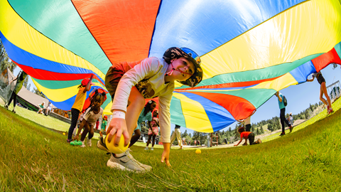 A child running joyfully beneath a large, colorful rainbow parachute during an outdoor summer camp activity at Biggie's Adventure Outpost at Big Bear Mountain Resort.