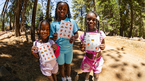 A happy group of children standing together outdoors under a shade structure, proudly holding up their colorful completed art projects at Biggie's Adventure Outpost at Big Bear Mountain Resort.