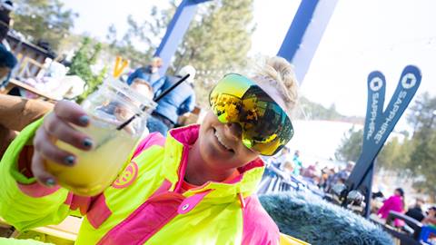 Adult in bright yellow and pink ski onesie cheersing the camera while smiling big.