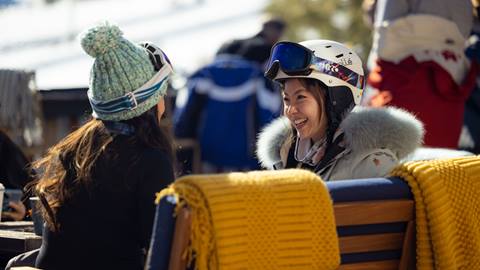Two adults in ski gear sitting at a fire pit mid discussion with two yellow blankets draped over chairs.