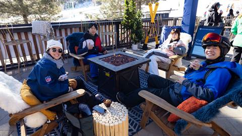 Adults in snow gear sitting in lounge chairs on sun deck around fire pit.
