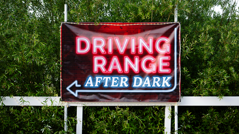 An outdoor daytime sign reading "Driving Range After Dark" with a left arrow, set against green trees at Big Bear Mountain Resort.