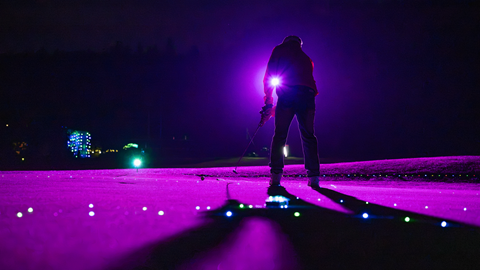 A person standing on the golf range turf at night under vibrant purple neon event lighting during Driving Range After Dark at Big Bear Mountain Resort.