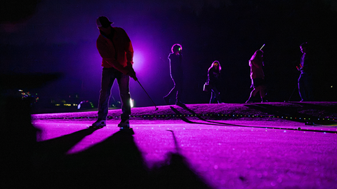 Silhouettes of people gathered on the grass turf under vibrant purple and pink neon lighting during the Driving Range After Dark event at Big Bear Mountain Resort.