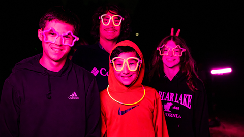 Three young people smiling and posing together under vibrant pink and purple neon lighting at Big Bear Mountain Resort's Driving Range After Dark event.