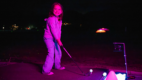 A person standing on the golf range grass illuminated by a vibrant pink and purple spotlight during the Driving Range After Dark event at Big Bear Mountain Resort.