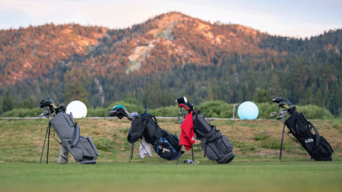 Daytime view of the golf driving range at Big Bear Mountain Resort featuring golf bags lined up on the grass with mountains and pine trees in the background.
