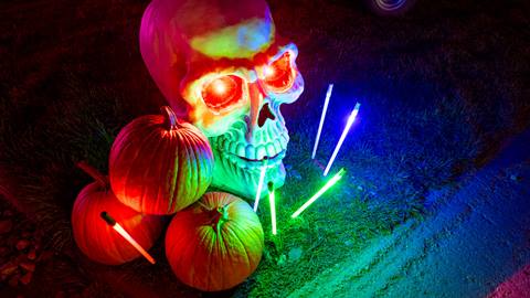 Three pumpkins on the grass laying next to a skull head with glowing red eyes and various color glow sticks glowing during the night.