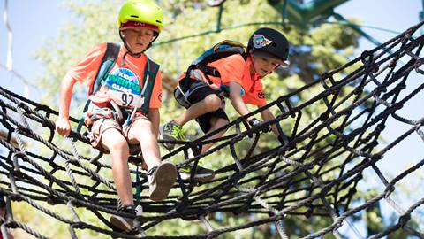Two kids in orange shirts crawling on a rope net obstacle course.
