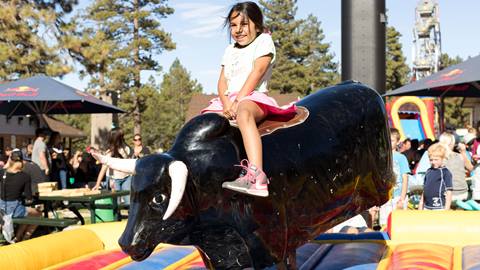 A kiddo, smiling very big wearing a green top, pink skirt, and pink shoes, riding a black mechanical bull during the summertime.