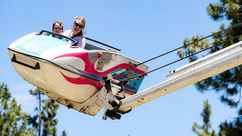 Adult and kid on a carnival ride with a blue sky and pine trees in background.