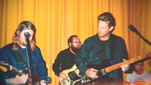 Three adults in dark clothing, all with guitars, performing a band set against a yellow backdrop.