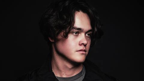 Close up side profile photo of an adult with dark hair, dark jacket with gray shirt underneath, and a silver chain around neck.