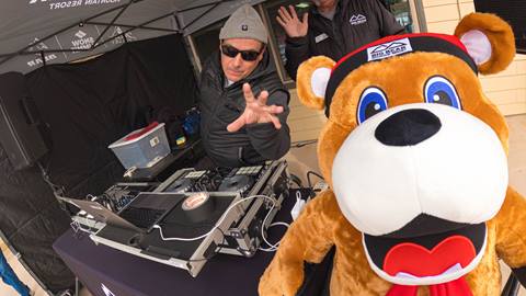 DJ Slipmatt behind his DJ setup, under a Big Bear Mountain Resort black eazeup tent, performing at a ski resort in the base area and standing next to an employee in a black jacket and mascot Biggie the Bear.