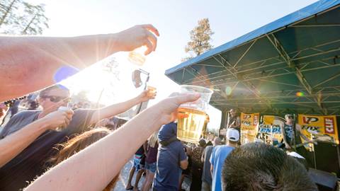 A live band performing to a crowd and the guests at the front are holding their beer cups up with the sun reflecting on them.