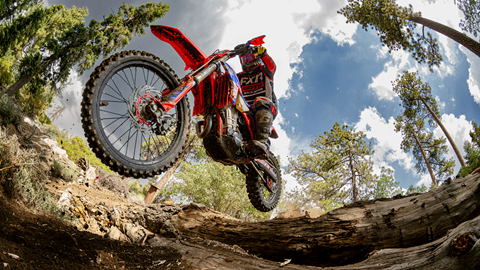 Motocross rider on red bike jumping over logs with clouds and sunny skies up above.