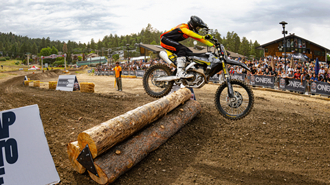 Motocross rider in dark with yellow jersey, jumping over logs on a dirt course in the base area of Bear Mountain, with clouds and sunny skies up above and a crowd cheering from the sidelines.