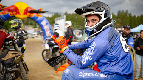 A motocross rider, in a black full face helmet and blue jersey and pant combo, on their bike throwing a hang loose shaka sign at the start gate for the event.