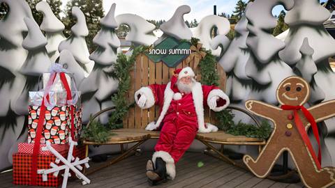Santa Claus at Snow Summit's Santa Comes to Summit event sitting on a Snow Summit green and brown wood bench with snowy trees, a candyman, and present cut outs.