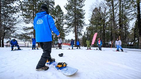 A Big Bear Mountain Resort snowboarder instructor on their snowboard looking at a group of snowboarders at the base area for lessons